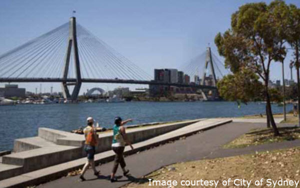 People walk along a waterfront path beside Blackwattle Bay, with a large cable-stayed bridge and city skyline visible in the background.