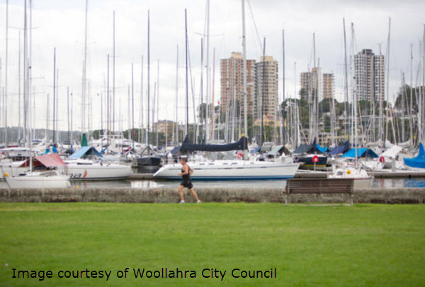 A grassy waterfront park overlooks a marina filled with sailboats and city buildings in the background, with a caption crediting the image to Woollahra City Council.