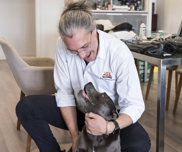A veterinarian kneels indoors while gently holding and interacting with a happy black dog.