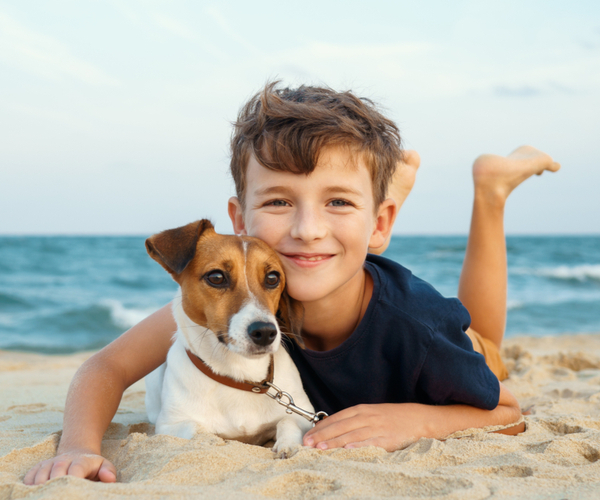A smiling boy lies on the beach beside a small dog, with the ocean in the background.