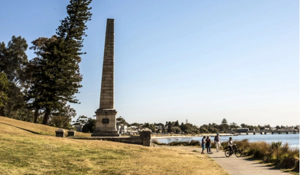 Burrawang Walk Kurnell in Botany Bay Council. Tall stone obelisk monument along a lake in a grassy waterfront park, with a paved path and a few people biking nearby.