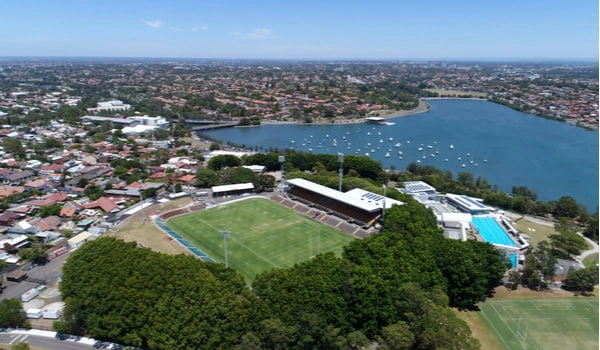 Aerial view of Leichhardt Oval in Sydney's Inner West. Oval sits beside the water dotted with boats and surrounded by houses and trees.