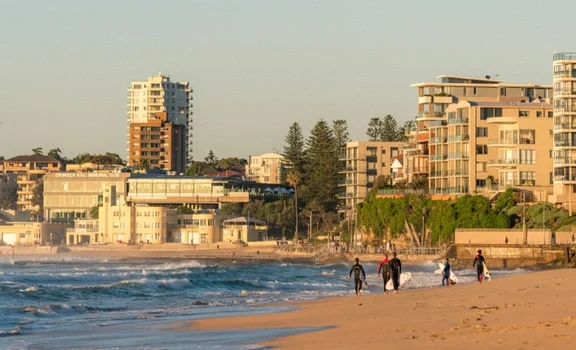 Sunlit Bondi beach in Lower North Shore with a few people carrying surfboards along the shore, with apartment buildings behind them.