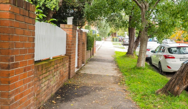 Randwick street sidewalk along a residential street with a brick wall and white fence on the left, and trees and parked cars on the right.
