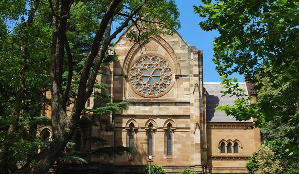 Stone church facade located in Woollahra. With a large circular rose window and arched windows, partially shaded by trees on a sunny day.